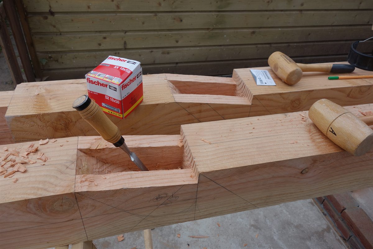 Twee balken op schragen met uitgestoken pengaten; er staat een beitel in het hout, ernaast liggen houten hamers, potloodlijnen en krullen.