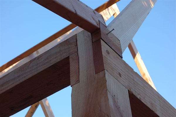 Van onderaf gefotografeerde verbinding boven op een houten stijl, waar meerdere balken en een schoor samenkomen; de deuvels zitten strak in het hout.