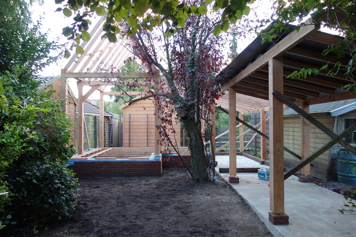 Overzicht in de tuin: links het houtskelet van het tuinhuis op een bakstenen plint, rechts de open constructie van de fietsenberging die om een boom heen is gebouwd.