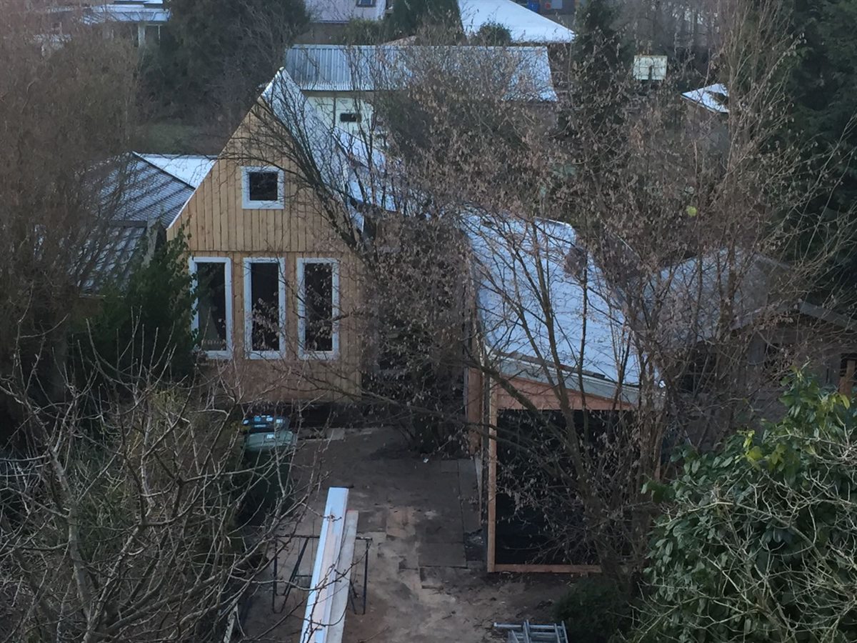 Van bovenaf gefotografeerd tuinhuis in de winter, met verticale houten gevelbekleding, witte kozijnen en een golfplaten dak, tussen kale takken.