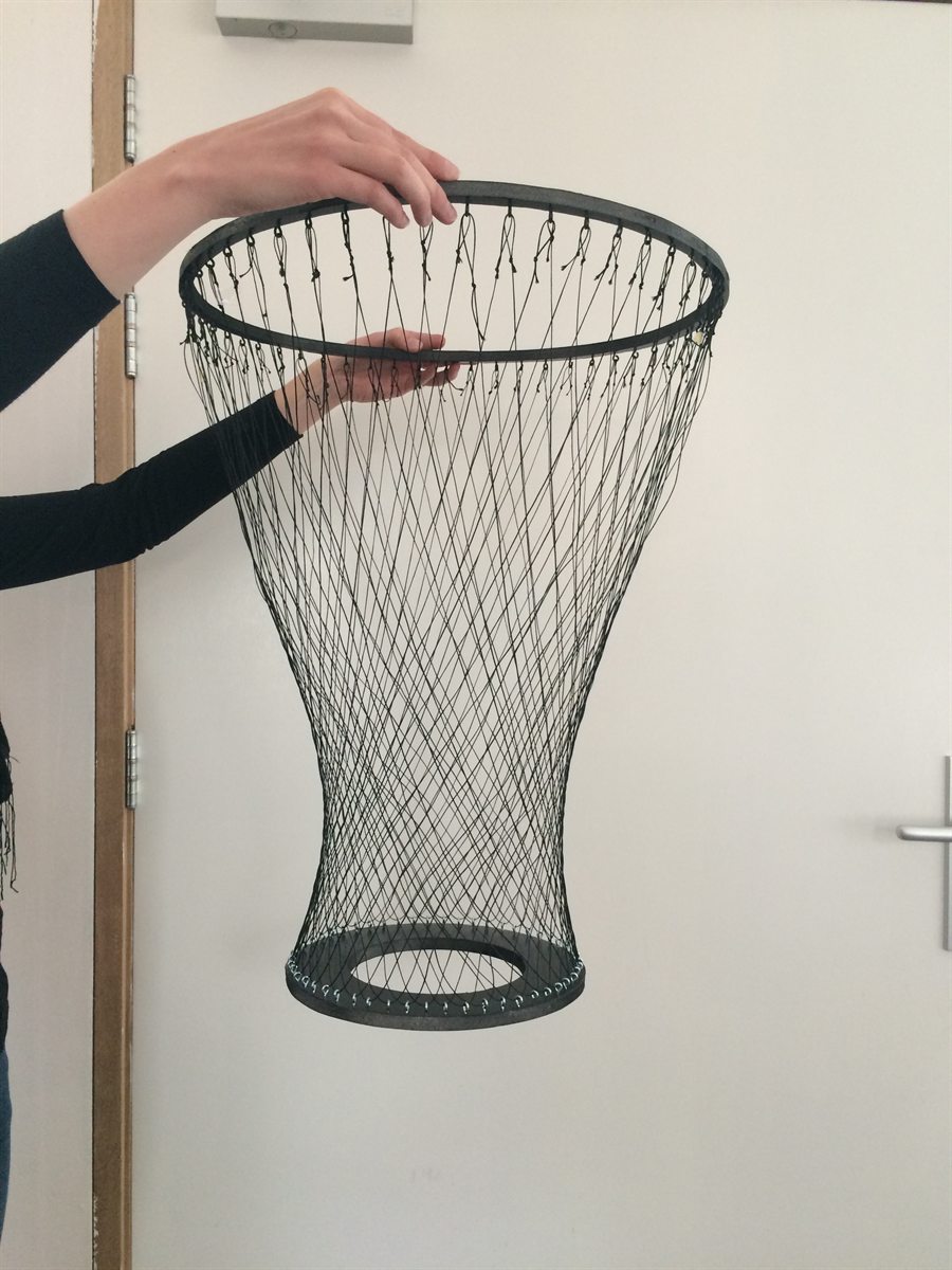 A hand holds up a model of the dreamcatcher: two steel rings with a finely knotted net of black thread between them, against a white door.