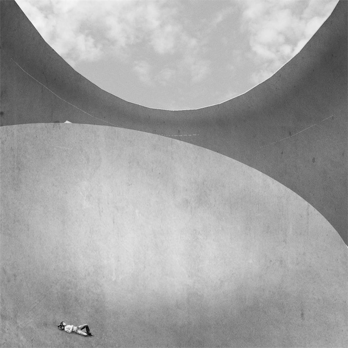 Someone lies alone on the curved concrete floor of the round shell; above them the opening cuts a sharp circle out of the clouded sky.