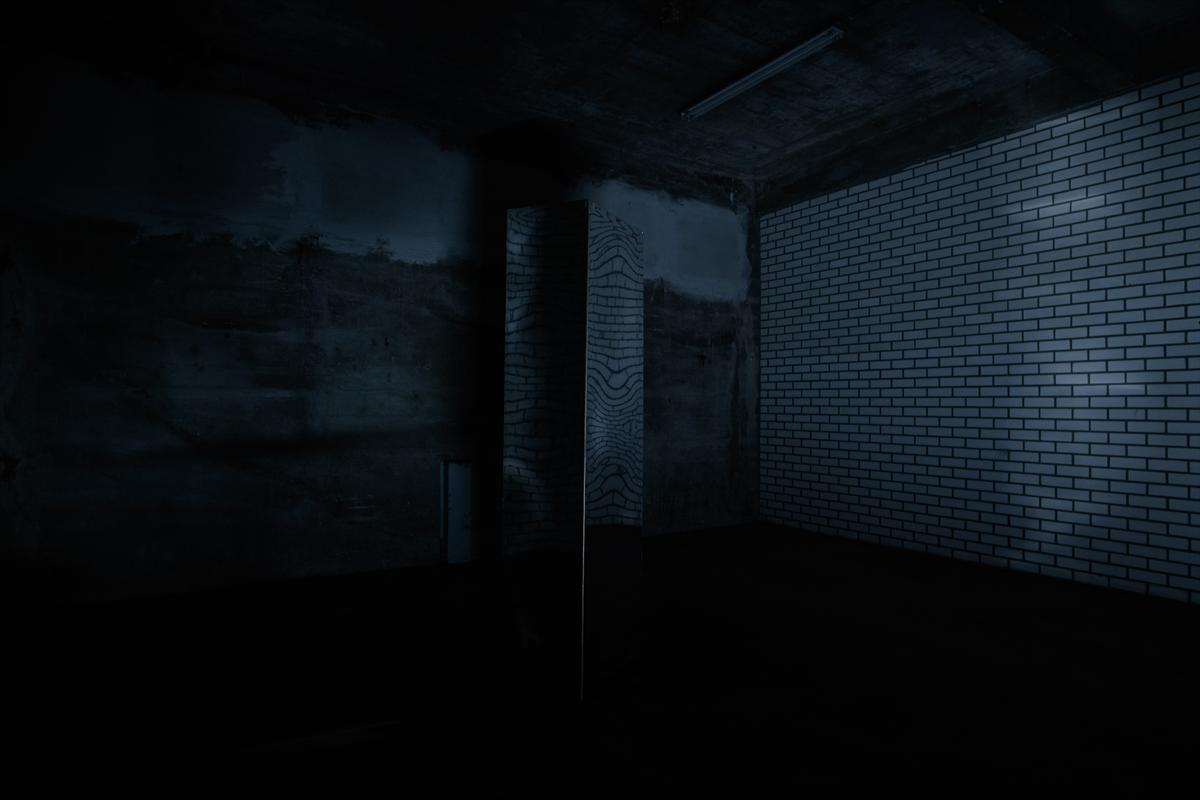 Almost entirely dark room in the bunker with a white tiled wall on the right, a concrete column in the middle and a faintly glowing projection on the back wall.