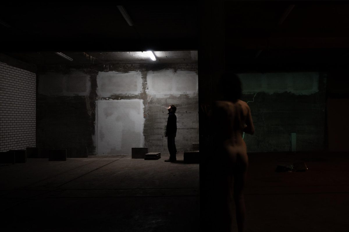 Two adjoining bunker spaces; on the left a figure stands under a fluorescent tube against a concrete wall, on the right a projected figure moves along a green line of light.