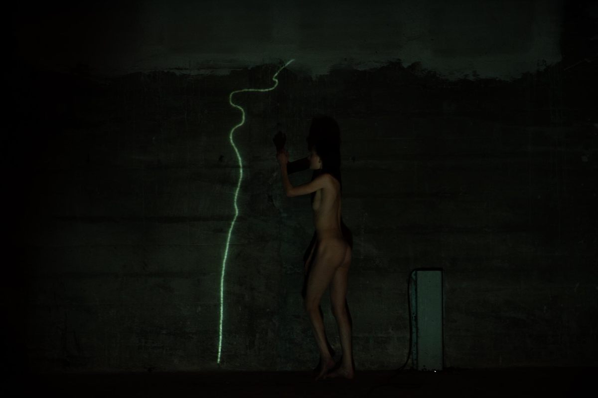 Almost entirely dark concrete space in which a nude figure with a raised arm leaves a glowing line on the wall.