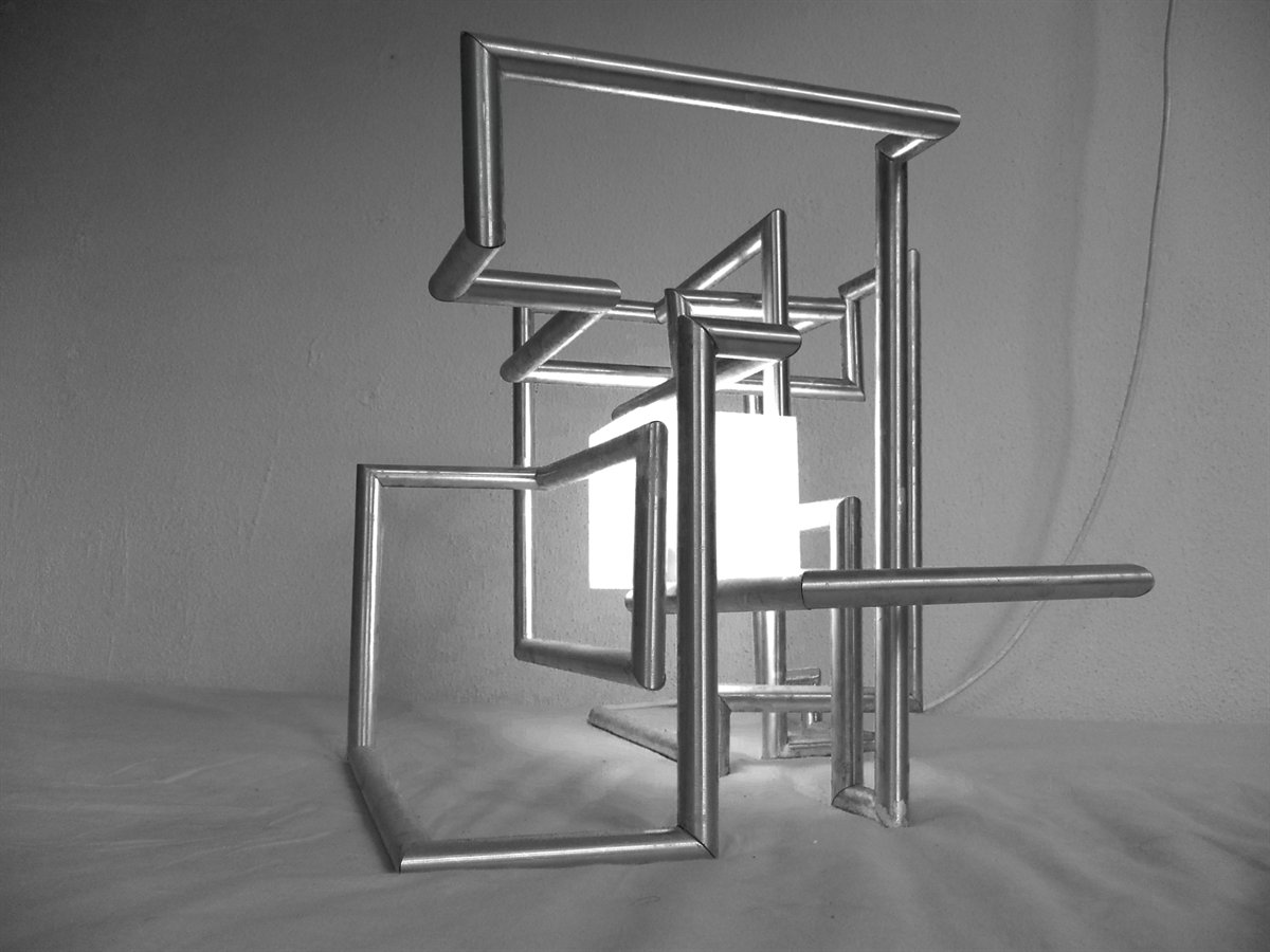 Zwart-witfoto van de lamp: aluminium buizen die als in elkaar grijpende rechthoeken door elkaar steken, met in het midden een oplichtende witte bak.