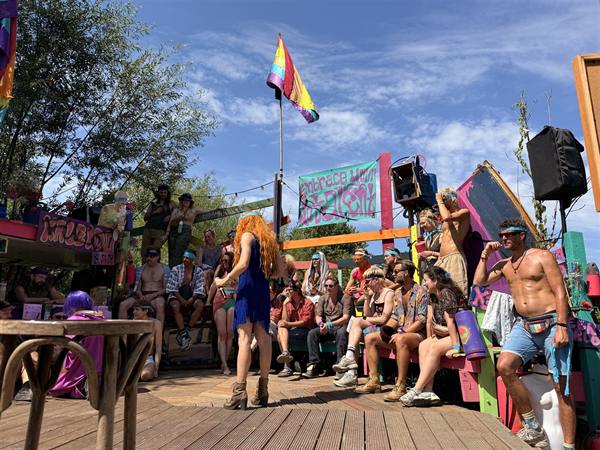 Het houten dek van de Queer Stage tijdens een voorstelling: een artiest in een blauwe jurk, publiek op gekleurde banken rondom, regenboogvlaggen en de felgekleurde KALEIDO-bar.