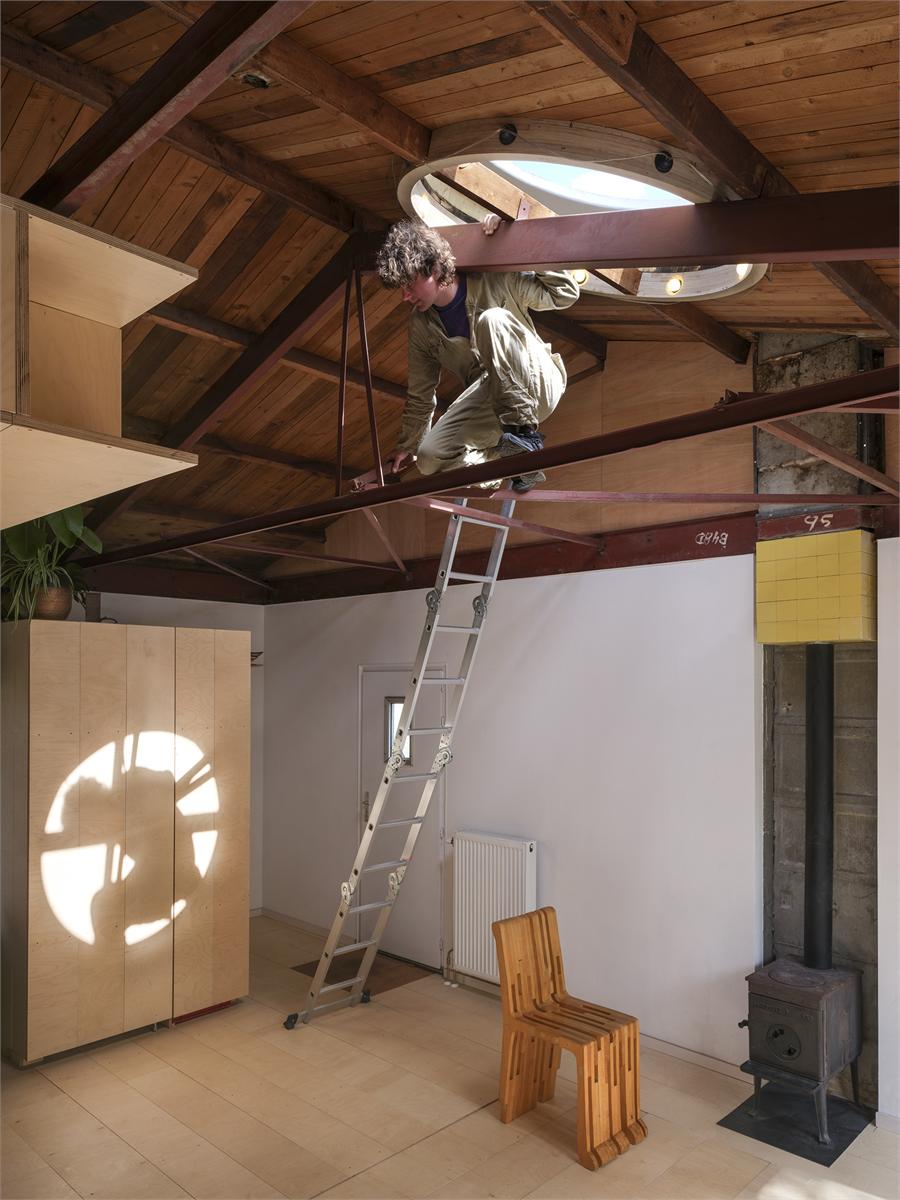 Someone climbs a ladder over a steel truss towards a round rooflight; on the left a birch plywood cupboard onto which the round window casts a patch of light, on the right a wood stove.
